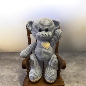 Könnte beinhalten: Ein handgehäkelter Teddybär in Hellblau mit einem cremefarbenen Herz, der in einem Holzschaukelstuhl sitzt. Der Bär hat schwarze Augen und ist handgefertigt. Die Arme und Beine des Bären sind so positioniert, als ob er sitzen würde.