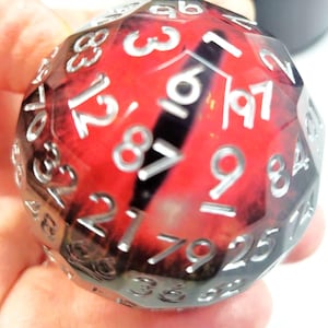 Puede incluir: Un dado poliédrico rojo y negro con números blancos. El dado se sostiene en una mano y tiene una superficie facetada. Los números son claramente visibles sobre el fondo rojo, con una franja negra.