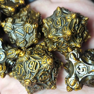 May include: A collection of intricately designed, metallic dice with a bronze and gold finish. The dice feature detailed patterns and numbers, suggesting a fantasy or tabletop gaming theme. The dice are held in a hand.