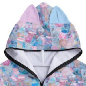 May include: A pink and blue hooded sweatshirt with a cat ear design. The sweatshirt has a colorful all-over print of various shapes and patterns.