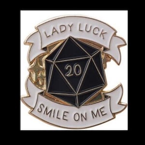 May include: A gold-coloured pin featuring a black twenty-sided die and a white banner that reads "Lady Luck Smile On Me".