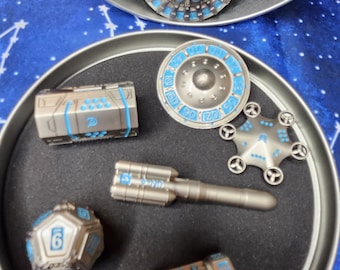 May include: A set of six silver and blue metal dice and tokens in a round tin container. The dice and tokens feature a variety of designs, including a spaceship, a rocket, and a cylinder.
