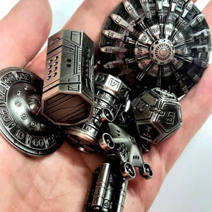 May include: A collection of silver-colored, intricately designed metal objects. The assortment includes a cylindrical piece, a multi-faceted die, a disc with numbers, and other mechanical-looking components. The objects have a detailed, industrial aesthetic.