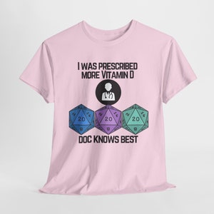 May include: Light pink t-shirt with the text "I WAS PRESCRIBED MORE VITAMIN D" above a graphic of a doctor and three colorful dice. The dice are blue, purple, and teal, each with the number 20 visible. The text "DOC KNOWS BEST" is below the dice.