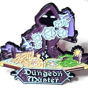May include: A purple and black enamel pin featuring a hooded figure holding dice. The pin has a landscape scene with a castle, river, and sun, with the words "Dungeon Master" in a banner.
