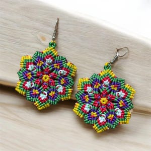 May include: A pair of handmade macrame earrings. Each earring features a colorful, floral design with yellow, green, red, purple, and white threads. The earrings have silver-colored fish hook ear wires and are displayed on a light wooden surface.