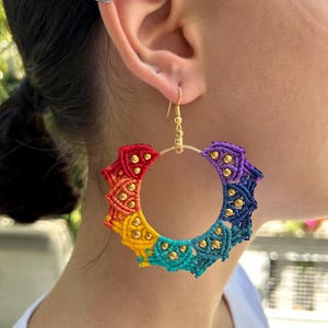 May include: Rainbow colored crocheted hoop earrings with gold beads and a gold hook. The earrings are hanging from a person's ear.
