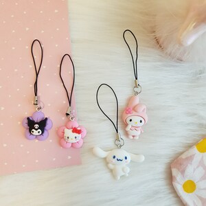 Op de afbeelding: Vier kleurrijke karakter-telefooncharms met zwarte touwlussen. De charms zijn een paarse bloem met een zwart kattengezicht, een roze bloem met een roze Hello Kitty-gezicht, een roze konijn met een witte jurk en een witte hond met blauwe ogen.