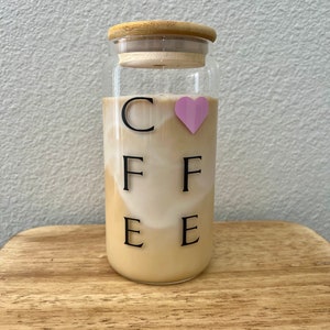 May include: A glass jar with a wooden lid and a pink heart sticker. The jar is filled with a light brown liquid and has the word "COFFEE" written on it in black letters.