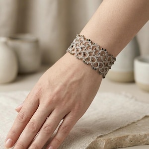 Könnte beinhalten: Ein breites, beigefarbenes Spitzenarmband mit einem komplizierten, bogenförmigen Schiffchenspitzenmuster. Die feine Fadenarbeit ist mit kleinen, dunklen, metallisch glänzenden Perlen verziert, die in das gewebte Design eingearbeitet sind.