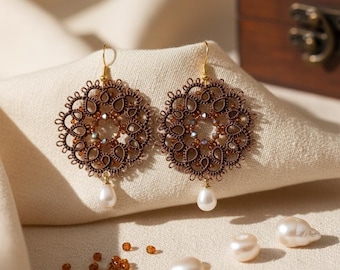 May include: A pair of round, brown tatting lace earrings featuring intricate thread work, small amber-coloured beads, and a single white teardrop pearl dangling from the bottom of each piece, suspended by gold-toned metal earring hooks.