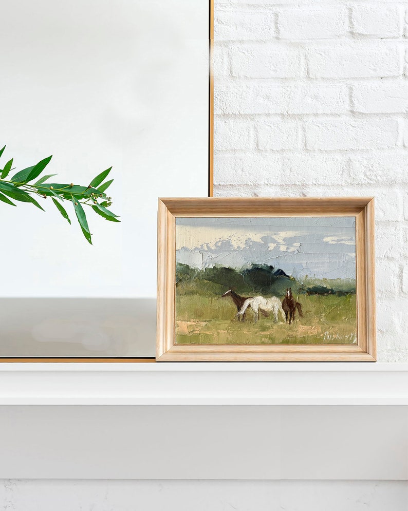 Puede incluir: Una pintura enmarcada de un paisaje con dos caballos en un campo. Los caballos son marr&oacute;n y blanco, y el campo es verde con un cielo azul en el fondo.