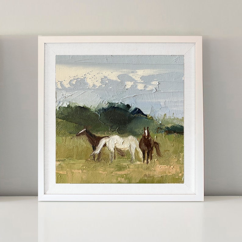 Puede incluir: Una pintura al &oacute;leo enmarcada de dos caballos de pie en un campo con una cordillera de monta&ntilde;as en el fondo. El cielo es de un azul claro con nubes blancas.