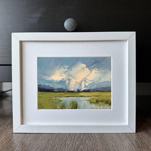 K&ouml;nnte beinhalten: Gerahmtes Landschaftsgem&auml;lde mit wei&szlig;em Rahmen. Das Kunstwerk zeigt ein Feld aus gr&uuml;nem und gelbem Gras, ein Gew&auml;sser und einen blauen Himmel mit wei&szlig;en Wolken. Das Gem&auml;lde ist ein Originalkunstwerk.
