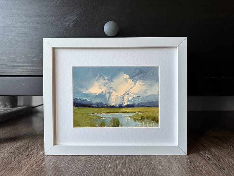 K&ouml;nnte beinhalten: Gerahmtes Landschaftsgem&auml;lde mit wei&szlig;em Rahmen. Das Kunstwerk zeigt ein Feld aus gr&uuml;nem und gelbem Gras, ein Gew&auml;sser und einen blauen Himmel mit wei&szlig;en Wolken. Das Gem&auml;lde ist ein Originalkunstwerk.