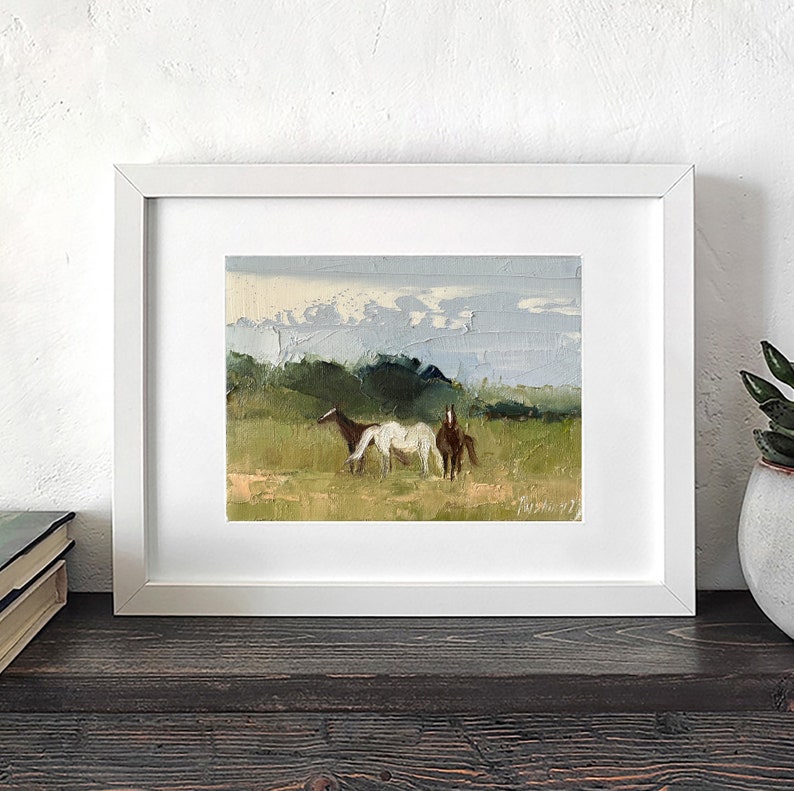 Puede incluir: Una pintura enmarcada de tres caballos en un campo. Los caballos son marrones y blancos, y el campo es verde. El cielo es azul y nublado. La pintura est&aacute; en un marco blanco.