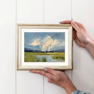 K&ouml;nnte beinhalten: Originales Landschafts-&Ouml;lgem&auml;lde in einem goldenen Rahmen. Das Kunstwerk zeigt eine lebendige Szene mit blauem Himmel, wei&szlig;en Wolken, gr&uuml;nen Feldern und einem Gew&auml;sser. Das Gem&auml;lde wird vor einem wei&szlig;en Hintergrund gehalten.