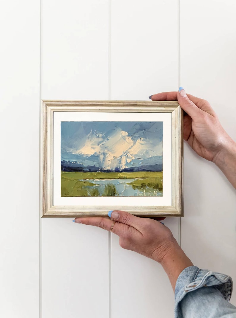 K&ouml;nnte beinhalten: Originales Landschafts-&Ouml;lgem&auml;lde in einem goldenen Rahmen. Das Kunstwerk zeigt eine lebendige Szene mit blauem Himmel, wei&szlig;en Wolken, gr&uuml;nen Feldern und einem Gew&auml;sser. Das Gem&auml;lde wird vor einem wei&szlig;en Hintergrund gehalten.