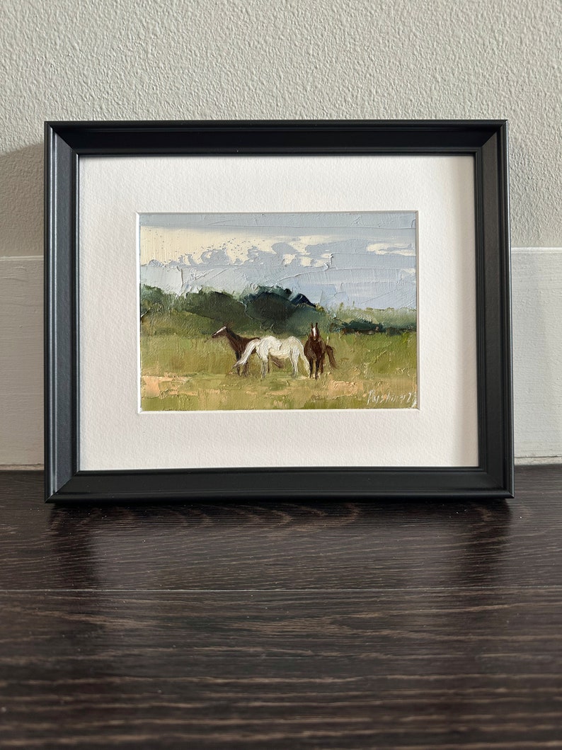 Puede incluir: Una pintura enmarcada de tres caballos en un campo. Los caballos son marrones y blancos, y el campo es verde. El cielo es azul y nublado.