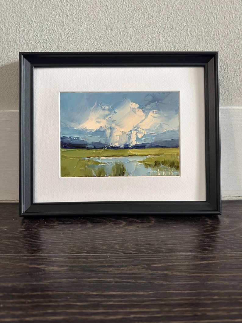 K&ouml;nnte beinhalten: Gerahmtes Landschafts-&Ouml;lgem&auml;lde mit einem lebendigen Himmel mit wei&szlig;en Wolken und einem gr&uuml;nen Feld. Das Kunstwerk ist in einem schwarzen Rahmen mit wei&szlig;em Passepartout gefasst, was einen auff&auml;lligen Kontrast erzeugt. Das Gem&auml;lde zeigt eine ruhige Szene im Freien.