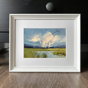 K&ouml;nnte beinhalten: Gerahmtes Landschaftsgem&auml;lde mit wei&szlig;em Passepartout und Rahmen. Das Kunstwerk zeigt eine lebendige Szene mit blauem Himmel, flauschigen wei&szlig;en Wolken und einem gr&uuml;nen Feld mit einem Gew&auml;sser. Das Gem&auml;lde hat einen strukturierten, impressionistischen Stil.