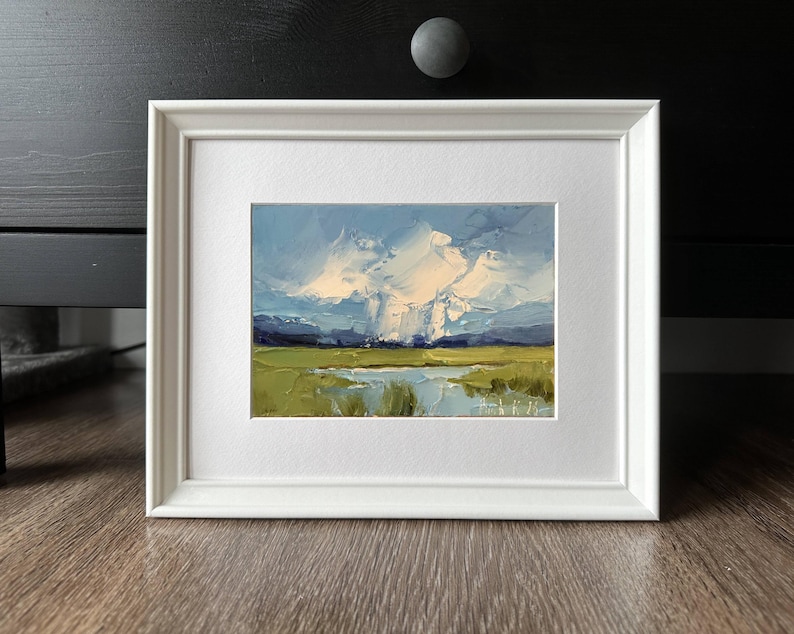 K&ouml;nnte beinhalten: Gerahmtes Landschaftsgem&auml;lde mit wei&szlig;em Passepartout und Rahmen. Das Kunstwerk zeigt eine lebendige Szene mit blauem Himmel, flauschigen wei&szlig;en Wolken und einem gr&uuml;nen Feld mit einem Gew&auml;sser. Das Gem&auml;lde hat einen strukturierten, impressionistischen Stil.
