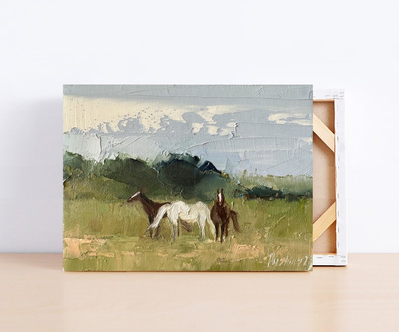 Puede incluir: Una pintura de tres caballos en un campo con una cordillera de monta&ntilde;as en el fondo. Los caballos son marr&oacute;n, blanco y marr&oacute;n. El cielo es de un azul claro con nubes blancas.