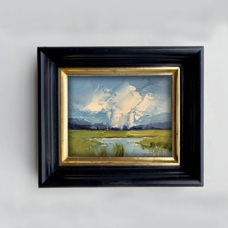K&ouml;nnte beinhalten: &Ouml;lgem&auml;lde einer Landschaftsszene, gerahmt in einem schwarzen Holzrahmen mit einem goldenen Innenrand. Das Kunstwerk zeigt einen bew&ouml;lkten Himmel &uuml;ber einem gr&uuml;nen Feld und einem Gew&auml;sser mit Bergen im Hintergrund.