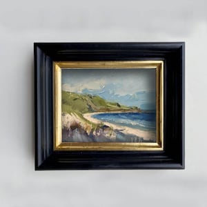 May include: An oil painting of a coastal landscape featuring a sandy beach, deep blue ocean water, and green hills under a cloudy sky. The artwork is encased in a thick, black wooden frame with a thin, inner gold-colored border.