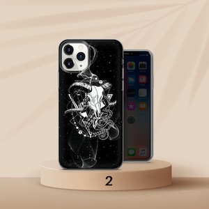 K&ouml;nnte beinhalten: Schwarze Handyh&uuml;lle mit wei&szlig;er Grafik eines Widder-Sch&auml;dels, geometrischen Formen und einem Oktopus. Die H&uuml;lle befindet sich auf einem beigen Podest neben einem Telefon mit beleuchtetem Bildschirm. Die Zahl "2" ist unten links aufgedruckt.