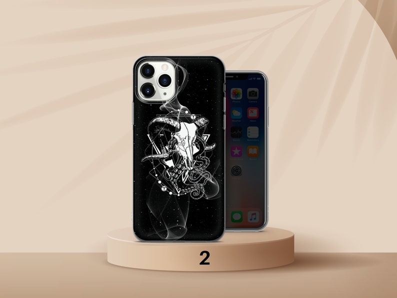 K&ouml;nnte beinhalten: Schwarze Handyh&uuml;lle mit wei&szlig;er Grafik eines Widder-Sch&auml;dels, geometrischen Formen und einem Oktopus. Die H&uuml;lle befindet sich auf einem beigen Podest neben einem Telefon mit beleuchtetem Bildschirm. Die Zahl "2" ist unten links aufgedruckt.
