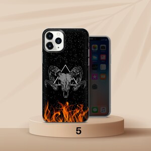 K&ouml;nnte beinhalten: Schwarze Handyh&uuml;lle mit einem Widder-Sch&auml;del-Design, einem wei&szlig;en Dreieck und einem Sternenhintergrund. Der Boden der H&uuml;lle zeigt orangefarbene Flammen. Ein weiteres Telefon ist rechts zu sehen.