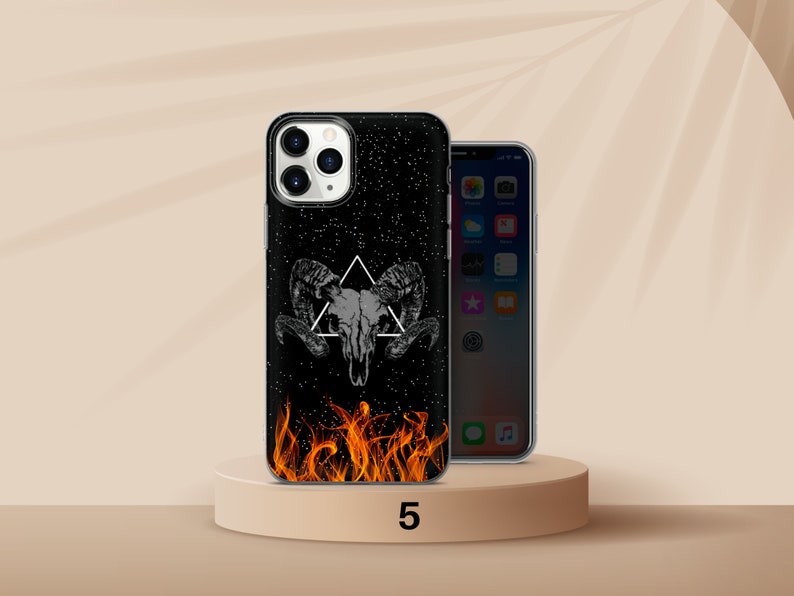 K&ouml;nnte beinhalten: Schwarze Handyh&uuml;lle mit einem Widder-Sch&auml;del-Design, einem wei&szlig;en Dreieck und einem Sternenhintergrund. Der Boden der H&uuml;lle zeigt orangefarbene Flammen. Ein weiteres Telefon ist rechts zu sehen.