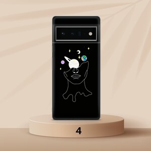 Puede incluir: Una funda de tel&eacute;fono negra con un contorno blanco de una cara con planetas y estrellas en los ojos. La funda de tel&eacute;fono est&aacute; sobre un pedestal marr&oacute;n claro con el n&uacute;mero 4 impreso en &eacute;l.