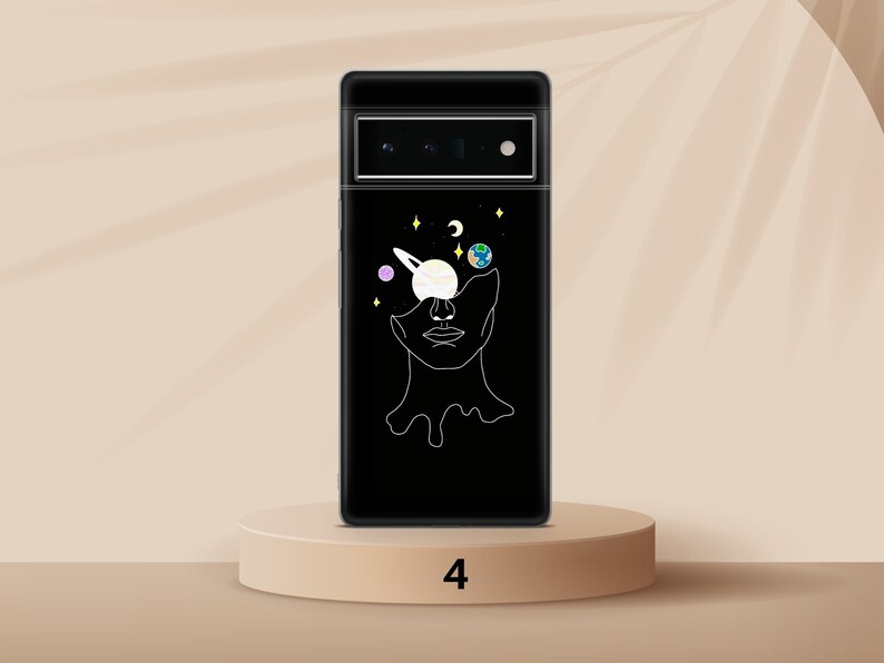 Puede incluir: Una funda de tel&eacute;fono negra con un contorno blanco de una cara con planetas y estrellas en los ojos. La funda de tel&eacute;fono est&aacute; sobre un pedestal marr&oacute;n claro con el n&uacute;mero 4 impreso en &eacute;l.