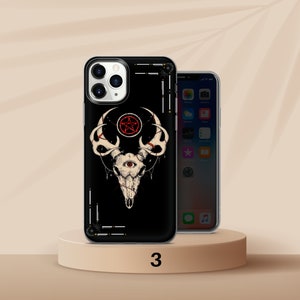K&ouml;nnte beinhalten: Schwarze Handyh&uuml;lle mit einem wei&szlig;en Sch&auml;deldesign, einem roten Pentagramm und einem Auge. Die H&uuml;lle befindet sich auf einem kleinen, runden, hellbraunen Sockel. Ein zweites Telefon ist rechts zu sehen.
