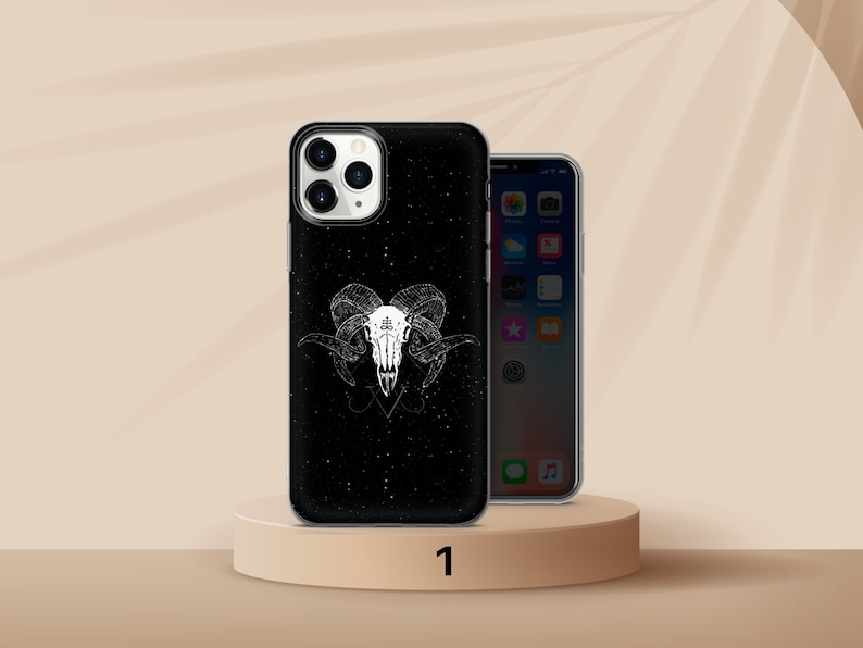 K&ouml;nnte beinhalten: Schwarze Handyh&uuml;lle mit wei&szlig;em Widder-Sch&auml;del-Design. Die H&uuml;lle hat einen gesprenkelten Hintergrund und eine detaillierte Illustration eines Widder-Sch&auml;dels mit H&ouml;rnern und einem Pentagramm-Symbol. Die Handyh&uuml;lle wird auf einem beigen Podest pr&auml;sentiert.