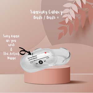 May include: A clear plastic case for Samsung Galaxy Buds or Buds+ with a customisable music player display. The display shows "Song Name" and "Artist Name" with a play/pause button and a timer. The case is on a pink surface.