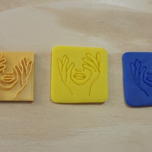 May include: Three square-shaped polymer clay stamps, each featuring a raised design of a face with hands covering the eyes. The stamps are yellow, yellow, and blue.