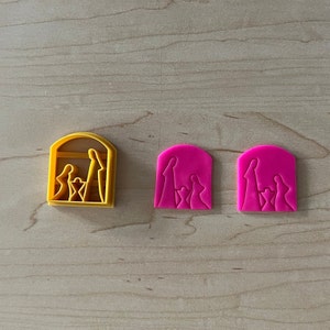 May include: Three cookie cutters, one yellow and two pink, shaped like the nativity scene with Mary, Joseph, and baby Jesus.