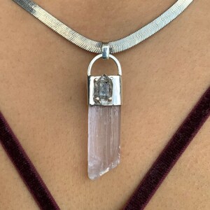 May include: A silver necklace with a pendant featuring a light pink crystal. The pendant is rectangular with a silver cap and a small, clear crystal on top. The necklace is a flat, silver chain.