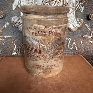 May include: A cylindrical beige ceramic jar featuring the text FABRIQUE DE CONFITURES FELIX POTIN and MAISONS DE VENTE 101 & 103 Boul. Sebastopol PARIS. The jar displays two circular illustrations of industrial factory buildings in brown and blue tones.
