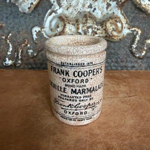 May include: A cylindrical ceramic jar with a cream-colored crackle glaze finish. The front features black printed text reading: ESTABLISHED 1874 FRANK COOPERS OXFORD SEVILLE MARMALADE HOME MADE WARRANTED PURE PREPARED ONLY BY Frank Cooper OXFORD REGISTERED TRADE MARK.