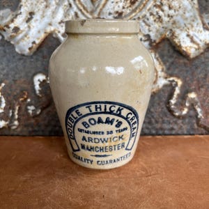May include: A small, off-white ceramic jar with a rounded body and a slightly flared rim. The jar features a blue and white label that reads "Double Thick Cream, Boam's, Ardwick, Manchester, Quality Guaranteed". The jar is likely vintage.