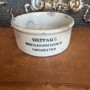 May include: A round, off-white ceramic pot with a visible crackled glaze finish. The exterior features black printed text that reads: SHIPPAM'S HIGH CLASS POTTED MEAT CHICHESTER. The container has a simple, open-top design with a smooth, rounded rim.
