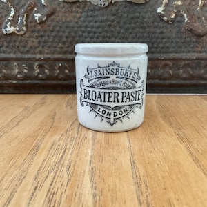 May include: Small, off-white ceramic jar with a lid. The jar features black text that reads "J. Sainsbury's Superior Home Made Bloater Paste London". The jar is sitting on a wooden surface.