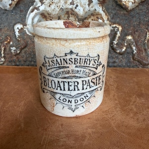 May include: A cylindrical ceramic pot with a cream-colored glaze and a network of fine brown cracks. The front features black printed text reading J. SAINSBURY'S SUPERIOR HOME MADE BLOATER PASTE LONDON, framed by decorative scrollwork.