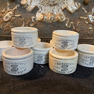 May include: A collection of small, off-white ceramic pots with the text "J. Sainsbury's Freshly Made Bloater Paste" in black lettering. The pots are cylindrical with a slightly flared rim, and some show signs of age and wear.