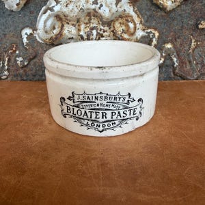 May include: A white ceramic pot with a cylindrical shape and a flat rim. The front features black printed text that reads J. SAINSBURY'S SUPERIOR HOME MADE BLOATER PASTE LONDON, framed by an ornate decorative scroll design with visible signs of wear.