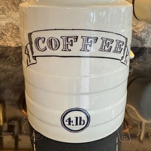 May include: A cream-colored ceramic canister with a lid, featuring a black hand-drawn style banner with the text COFFEE and a circular emblem below containing the text 4.lb. The cylindrical container has horizontal ridges along its body.